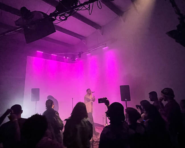 A performer sings on stage illuminated by purple lights while an audience watches from in front.