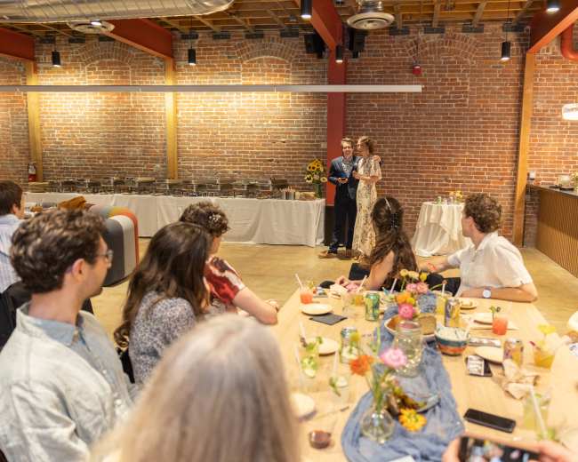 A couple stands at the front of a decorated event space while guests seated at a long table listen attentively.