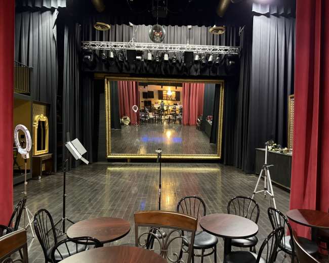 The image shows a stage with a large mirror framed in gold, surrounded by red curtains, and several tables and chairs set up in the foreground.
