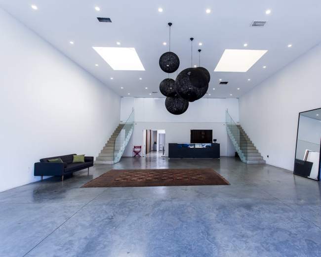 A spacious, minimalist interior features a large open area with concrete flooring, two sets of stairs, a dark rug, a modern sofa, and circular hanging light fixtures.