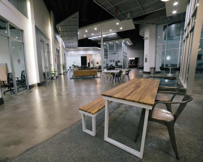 The image shows an open office space featuring a long wooden table with metal chairs, large glass windows, and various workstations in the background.