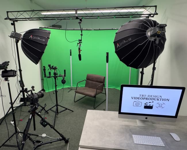 The image shows a video production setup with a green screen, lighting equipment, a camera on a tripod, a microphone, an empty chair, and a computer displaying a design screen.