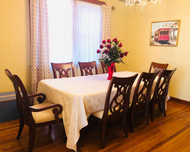 A dining room features a large table surrounded by eight wooden chairs, with a vase of roses on the table and a painting of a tram on the wall.