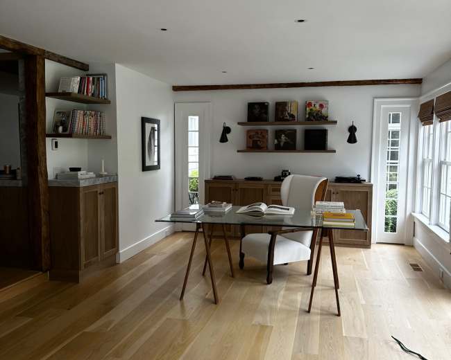 A modern, minimalistic home office features a round table with books, a white chair, and shelves in the background, all set on light wood flooring.