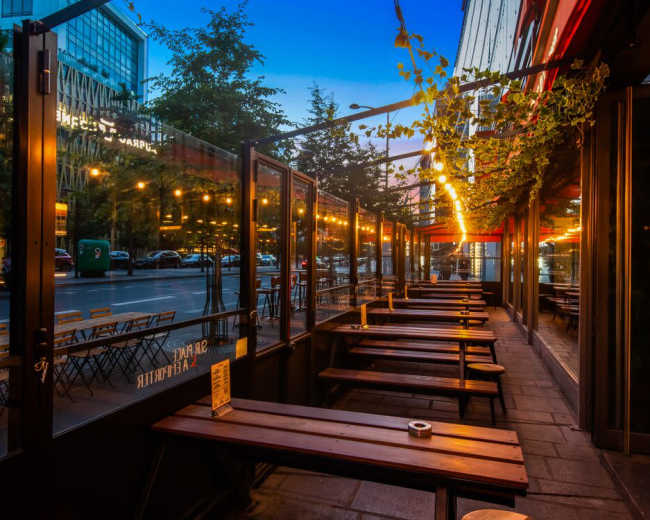 The image shows an outdoor dining area with wooden tables and benches, lined by glass panels, illuminated by string lights, and facing a street in an urban setting at dusk.