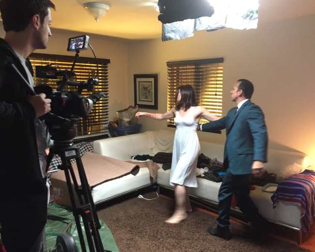 A woman in a white dress dances with a man in a suit while a cameraman records in a living room filled with scattered clothes and blankets.