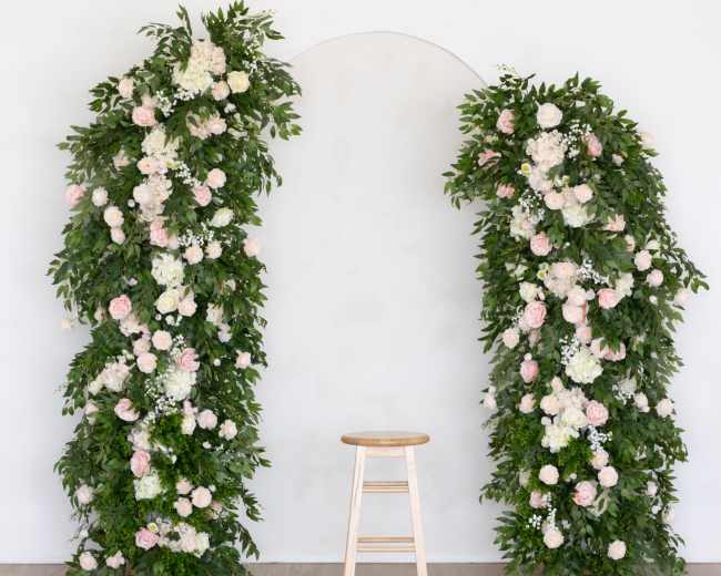 The image features a minimalist floral arch adorned with greenery and white and pink flowers, flanking a simple wooden stool against a white wall.