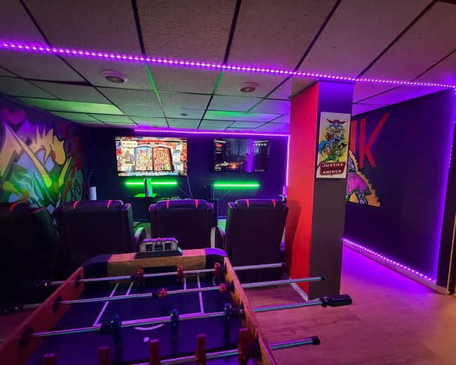 The image shows a brightly lit game room with foosball, comfortable seating, and two large screens displaying games, surrounded by colorful wall art.
