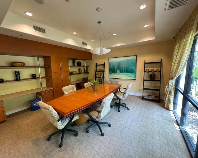 A conference room features a long wooden table surrounded by six chairs, with shelves displaying decorative items and a large artwork on the wall.