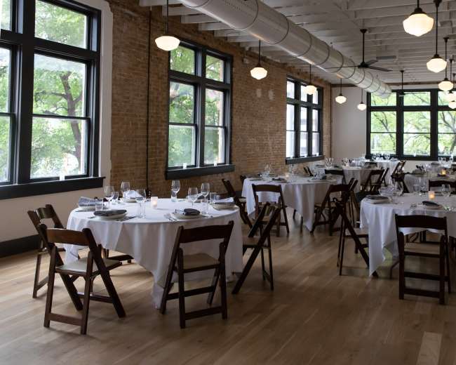 The image shows a dining room with several round tables set with white tablecloths and dinnerware, surrounded by large windows that allow natural light to enter.