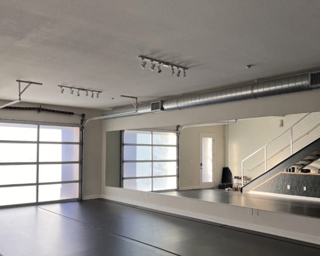 The image shows an empty gym or studio space with a mirrored wall and large glass garage door, illuminated by overhead lighting.