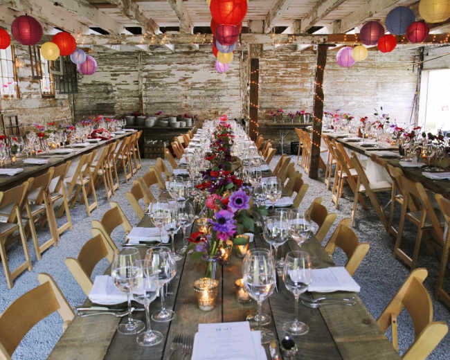 A long banquet table is set for dining with floral centerpieces and surrounded by wooden chairs in a rustic hall adorned with colorful lanterns.
