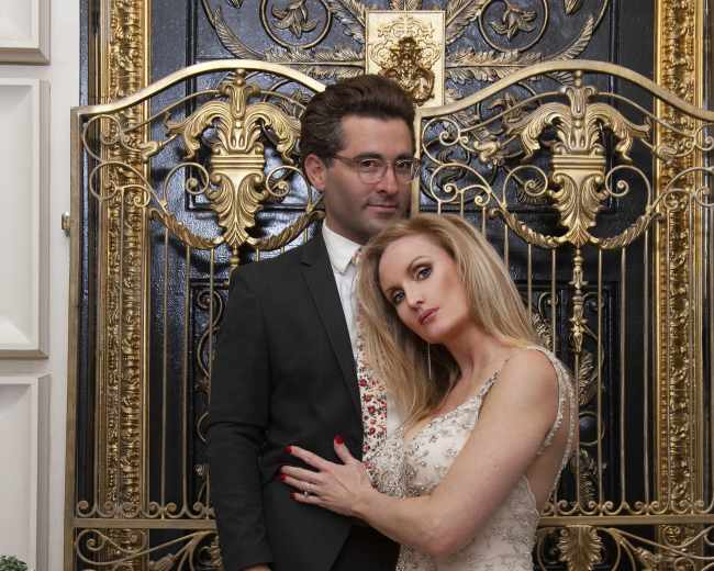 A couple stands together in formal attire in front of an ornate, gold-accented iron gate.