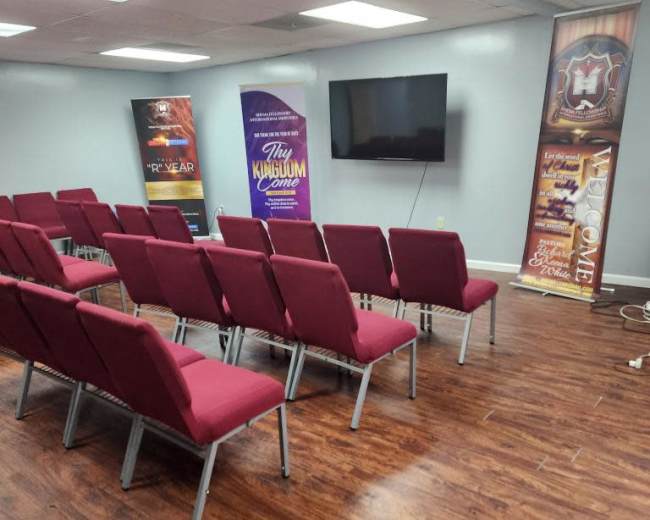 The image shows a meeting room with red chairs arranged in rows facing a blank television screen, surrounded by banners on the walls.