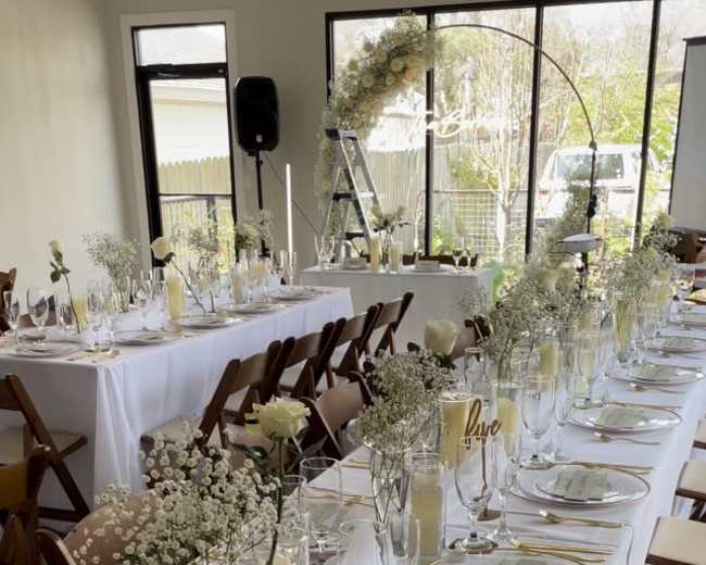 A long banquet table is set with elegant place settings, floral centerpieces, and candles, all arranged in a well-lit event space.