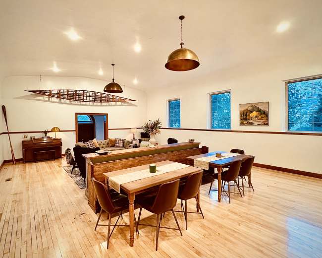 The image shows a spacious, well-lit dining and living area featuring a long wooden table, chairs, and decorative elements like a boat oar and artwork on the walls.