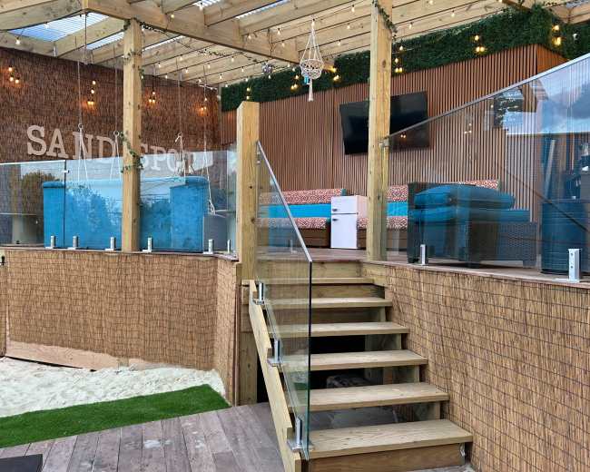 The image shows a wooden deck area featuring a staircase leading down to a sand-filled space, with a glass barrier and various seating arrangements under a partially covered structure.