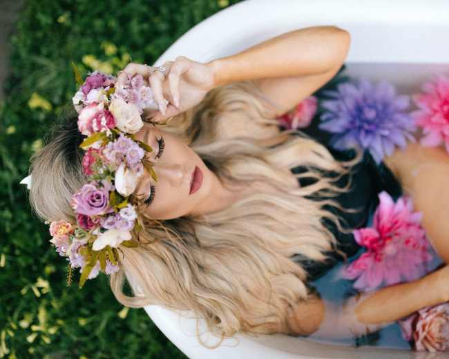 A woman with long hair wearing a floral crown relaxes in a bathtub filled with water and colorful flowers.