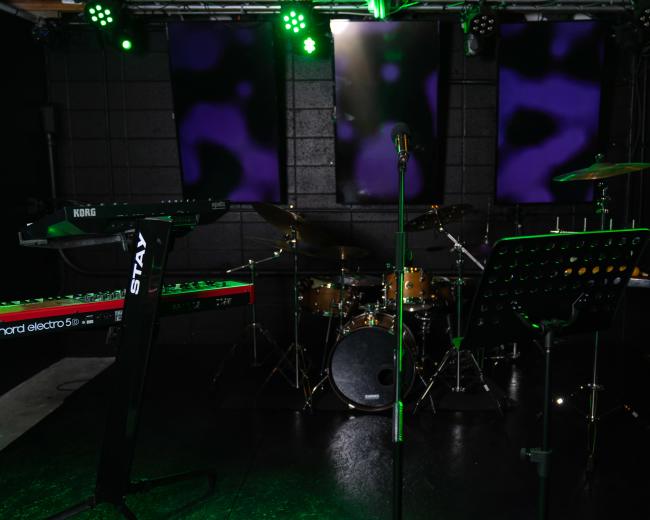 A music performance setup with a microphone in front of a drum kit, keyboard, and bass guitar, all under green stage lights and illuminated backdrops.