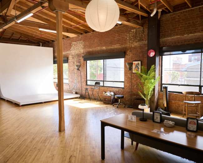 The image shows a spacious, well-lit studio with wooden flooring, large windows, a white backdrop on a raised platform, and a table with decorative items.