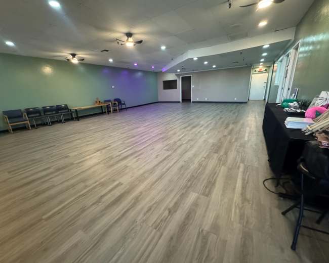 The image shows a spacious interior room with wooden flooring, green walls, and several ceiling fans, featuring a seating area along one side and a table covered with items on the right.