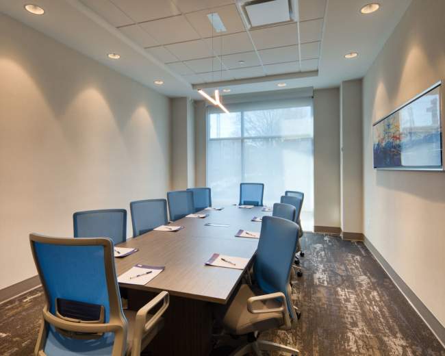 A modern conference room features a long table surrounded by eight blue chairs, with large windows letting in natural light and minimal wall decorations.