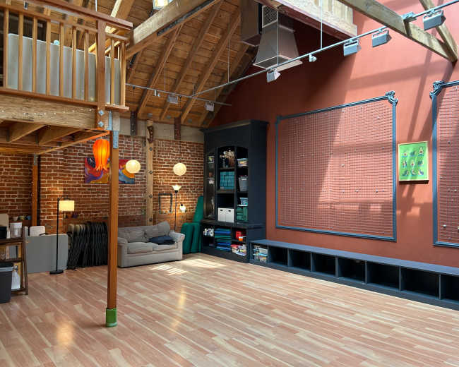 The image shows an expansive open space with wooden beams, a light hardwood floor, and a wall adorned with storage units and a framed green artwork.