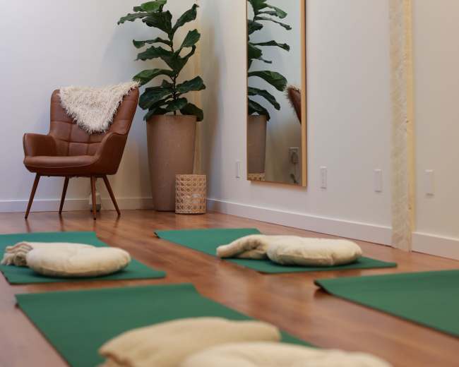 The image shows a serene room with green yoga mats laid on a wooden floor, a mirror on the wall, a potted plant, and a brown leather chair with a decorative throw.