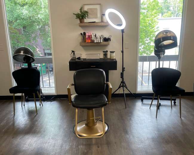 The image shows a modern hair salon setup with two black chairs, a styling chair in the center, a ring light, and two hooded dryers positioned against a wall of windows.
