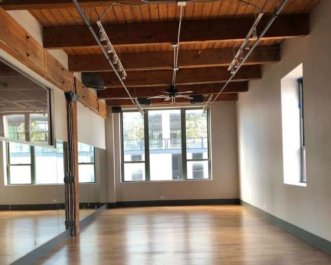The image shows an empty room with wooden beams, hardwood flooring, and large windows reflecting natural light.