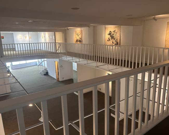 The image shows a multi-level art gallery with white railings and several artworks displayed on the walls.