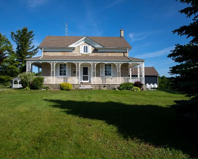 A two-story house with a wraparound porch sits amid a grassy landscape under a clear blue sky.
