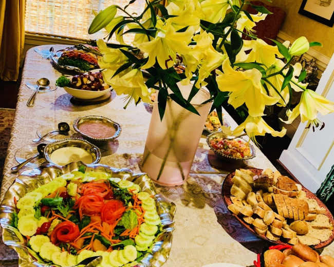 A dining table is set with a large salad, various side dishes, bread, and a vase of yellow lilies.