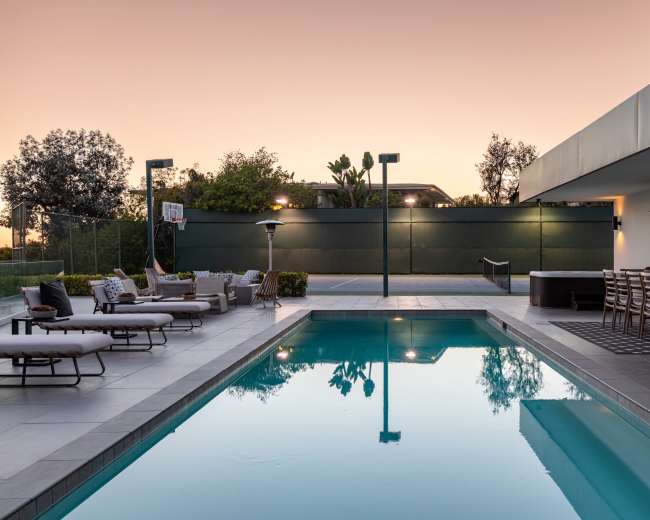 A modern pool area featuring loungers, a dining set, and a basketball hoop against a backdrop of greenery and a sunset sky.
