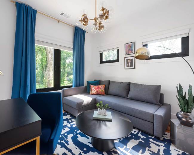 The image shows a modern living room featuring a gray sectional sofa, a round black coffee table, a blue and white patterned rug, and large windows with blue curtains.