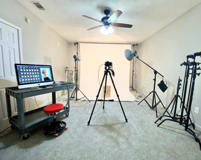 A photography studio is set up with a computer on a cart, a camera tripod, and multiple lights on stands facing a white backdrop.
