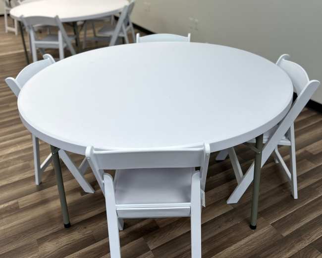 The image shows a room with several circular tables surrounded by white folding chairs, all set on a wooden floor.