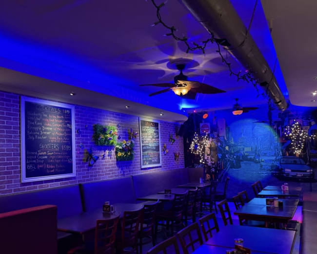 The image shows an interior view of a bar with purple lighting, brick walls, and a mural, featuring tables, chairs, and chalkboard menus displaying drinks.