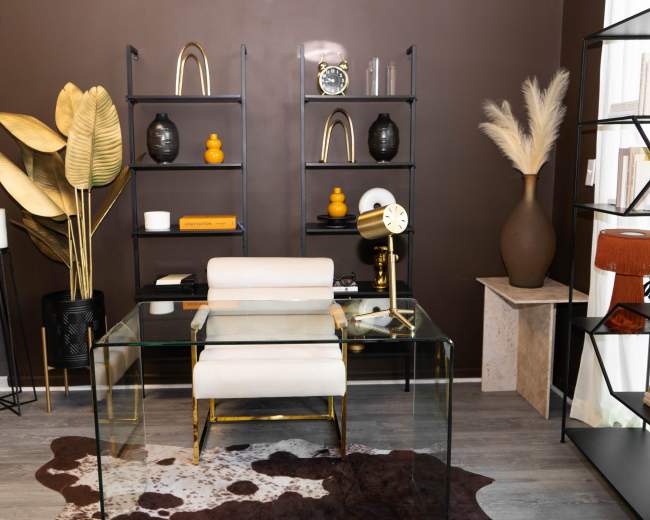 The image shows a modern office space featuring a glass desk, a white chair, and a variety of decorative items on black shelves against a brown wall.