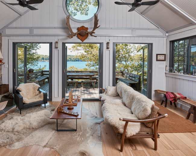 The image features a cozy living room with a stone fireplace, wooden furniture, and large windows overlooking a lakeside deck.