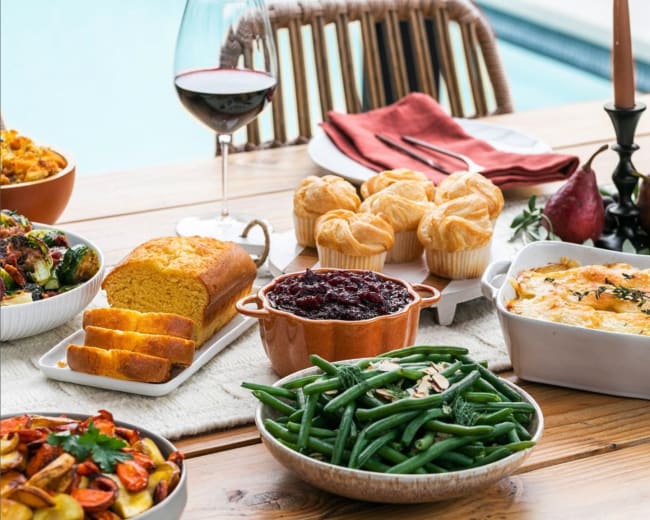 A wooden table is set with various dishes, including roasted vegetables, green beans, cornbread, cranberry sauce, muffins, and a creamy casserole, accompanied by a glass of red wine.