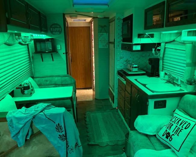 The image shows the interior of a camper, featuring a small kitchen area, a seating space, and a hallway leading to a bathroom or bedroom, all illuminated with green light.