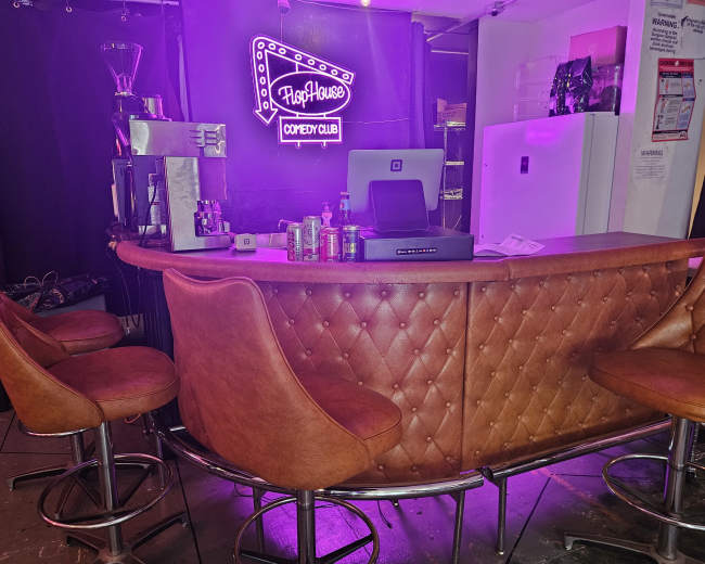 The image shows a cozy bar area with a brown leather counter, several high stools, and a purple neon sign that reads "FlopHouse Comedy Club."