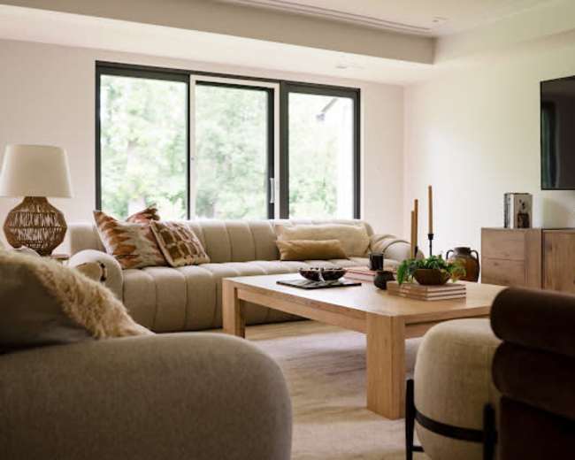 The image shows a modern living room with two sofas, a wooden coffee table, and large windows allowing natural light to enter the space.