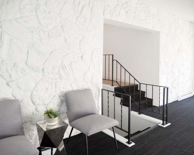 The image shows a minimalist interior with two gray chairs next to a small table, a coffee table, and a staircase leading to a higher level, all against a textured white stone wall.