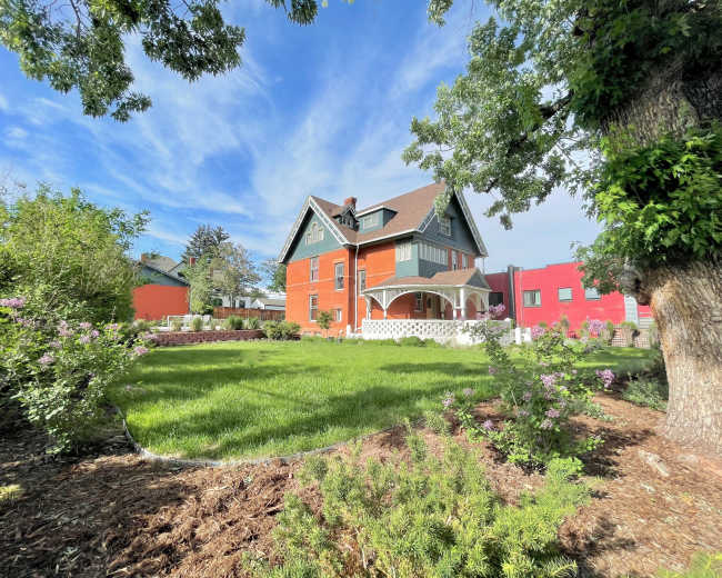 A large, two-story, red and green house sits in a spacious yard surrounded by trees and shrubs.