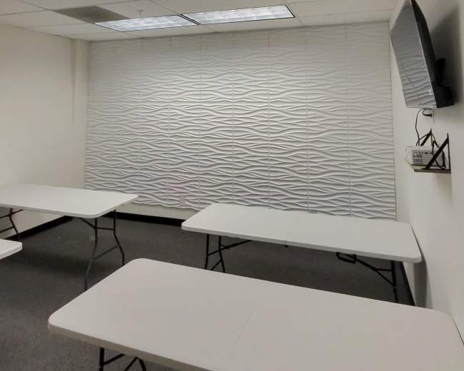 The image shows a modern classroom setting with four folding tables arranged and a textured white wall featuring a mounted television.