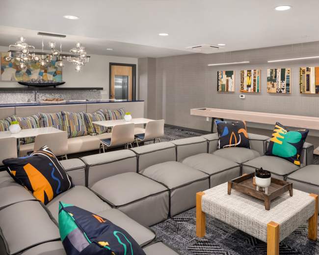 A modern communal lounge area featuring a sectional sofa with decorative pillows, a dining space with striped chairs, and a long table with wall-mounted artwork.