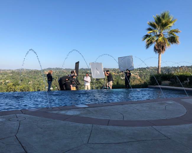 A group of people is working near a pool with fountains, using reflectors and lights against a backdrop of hills and a clear blue sky.