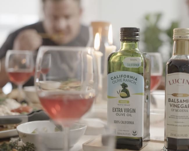 A bottle of California olive oil and a bottle of balsamic vinegar are placed on a dining table where people are enjoying a meal.